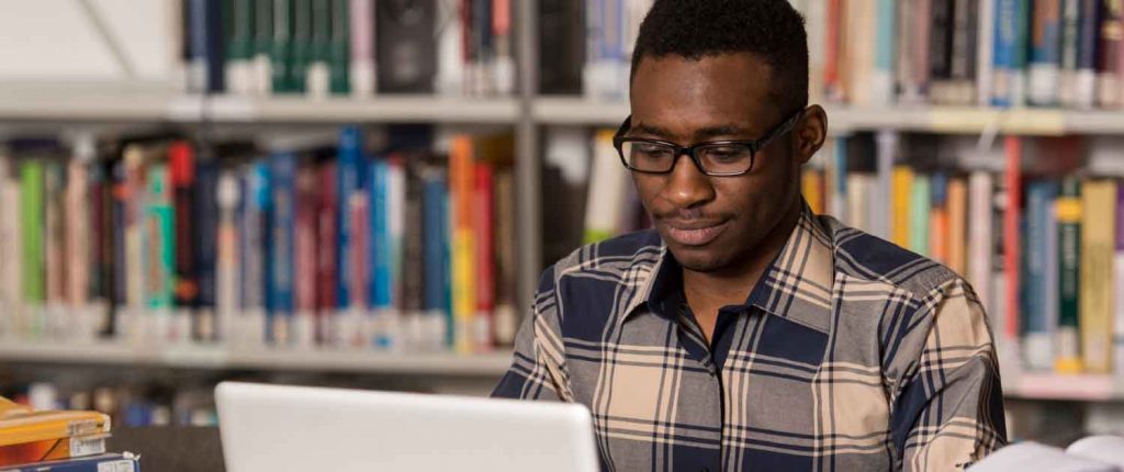 Black man wearing glasses and plaid shirt uses laptop computer. Library books are on a shelf behind him