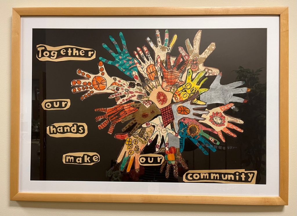 Framed art, black background with children's traced hands on paper decorated in unique ways. Words on art:Together our hands make our community.