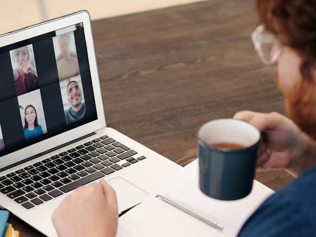Man wearing glasses and holding coffee mug looks at his laptop screen showing multiple faces. Zoom meeting