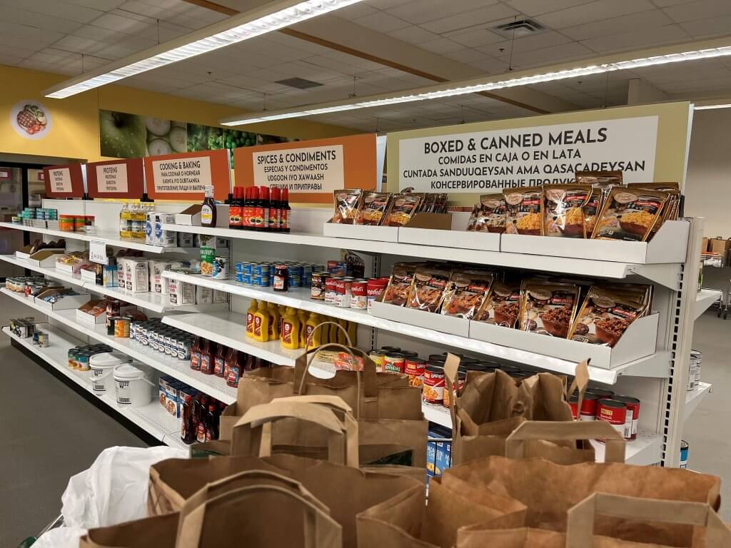 A Variety of Boxed and Canned Goods Line the Food Shelves at IOCP. Signage includes multi languages, e.g. Spanish, Somaili and Russian.
